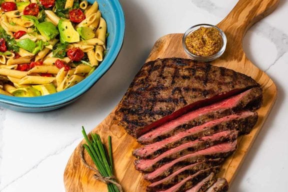 flank steak on a cutting board with a bowl of pasta salad