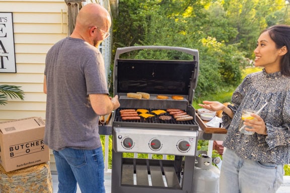 Two people with drinks standing by the grill as it cooks meat