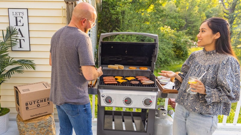 Two people with drinks standing by the grill as it cooks meat