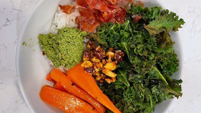 rice topped with roasted kale, carrots, proscuitto, hazelnuts, and basil pesto in a bowl