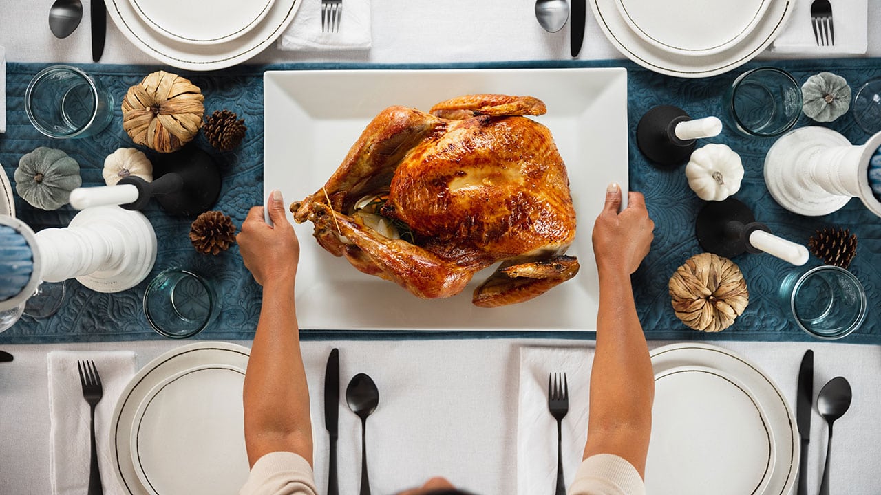 person placing a tray with a roasted turkey on a set dinner table