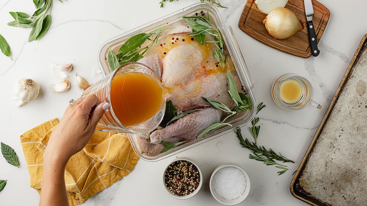 person pouring brine over a turkey in a dish with seasoning and herbs