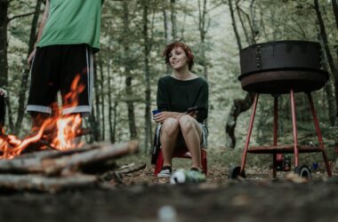 Woman sits by a campfire and a grill in the woods.