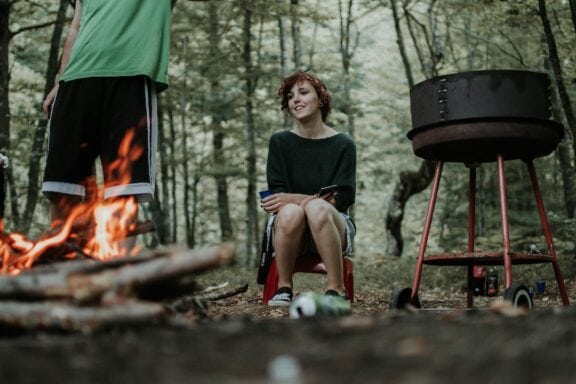 Woman sits by a campfire and a grill in the woods.