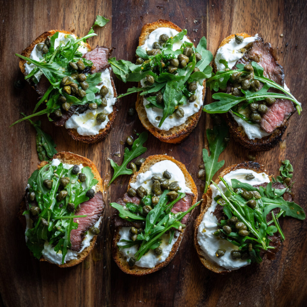 Filet Crostini with Horseradish Cream, Arugula and Capers on a chopping board